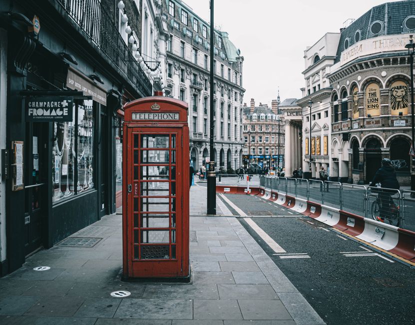 A traditional red British telephone box positioned on a pavement outside a commercial building in an urban street scene. The sidewalk features a storefront window with signage, and adjacent to the telephone booth, there are black bollards and a section of the road marked for traffic management. In the background, classic and modern architecture lines the street, with pedestrians and cyclists visible further along the pavement. The scene suggests an active city environment typical of London, within a context where house removals and furniture transport could be organized by Waterloo Man and Van as part of a home relocation or moving service, with the street ready for loading and transportation activities.