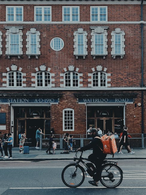 A man riding a bicycle with a large orange backpack on his back is moving along a street in front of a historic red-brick building with a sign reading Waterloo Station A.D. 1910 and Waterloo Road. The building features multiple large windows, some with white decorative stonework, and a small rectangular window near the center. On the sidewalk, several people are standing or walking, some with luggage or bags, indicating a busy urban environment. Inside the open entrances of the station, glimpses of interior activity can be seen. This scene depicts a typical day in Waterloo, with pedestrians and cyclists, emphasizing the area's transportation hub and the surrounding busy street scene connected to house and office relocations, as managed by Waterloo Man and Van in London.